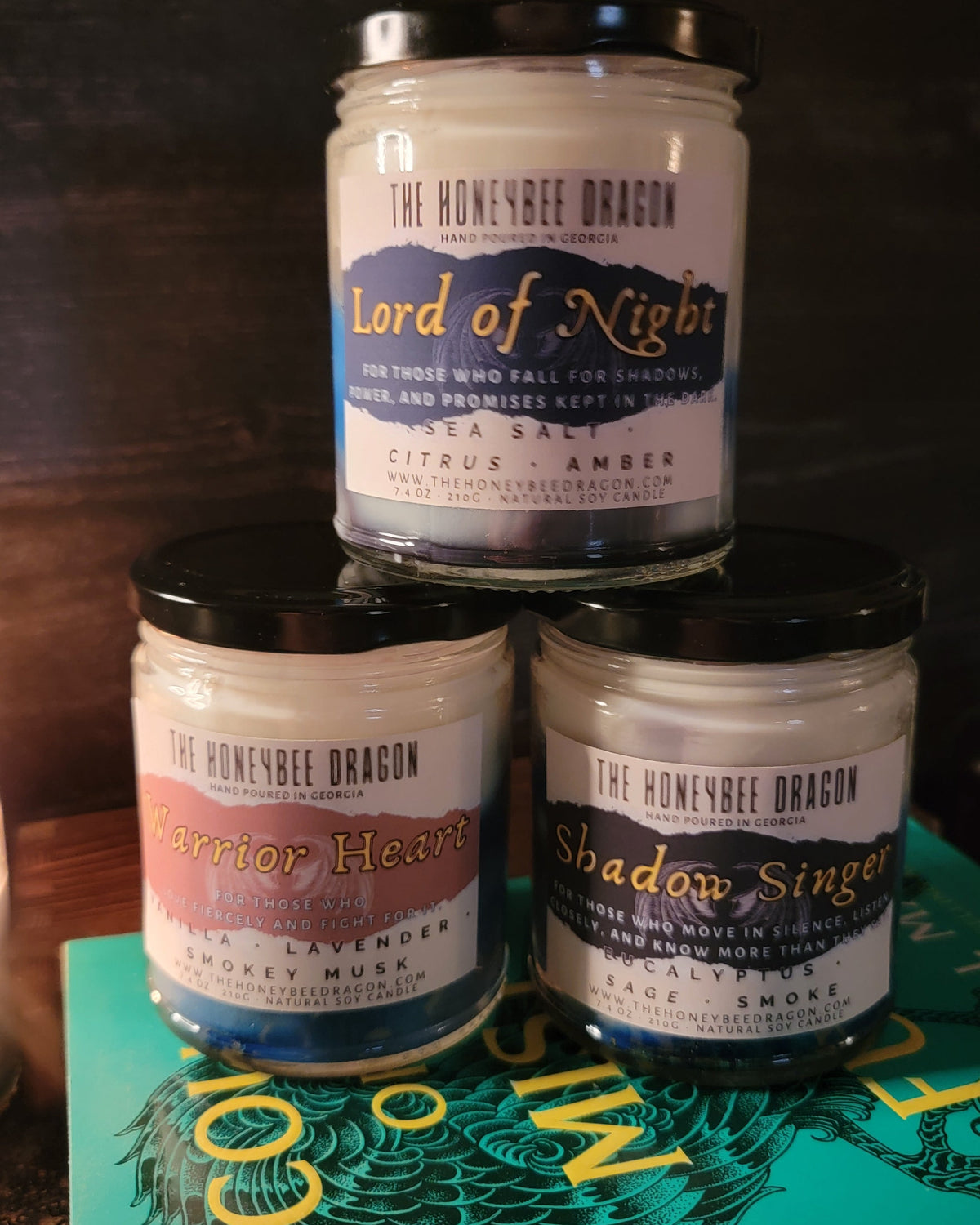Three stacked soy candles from The HoneyBee Dragon: Lord of Night, Warrior Heart, and Shadowsinger, displayed on a fantasy book against a dark background.