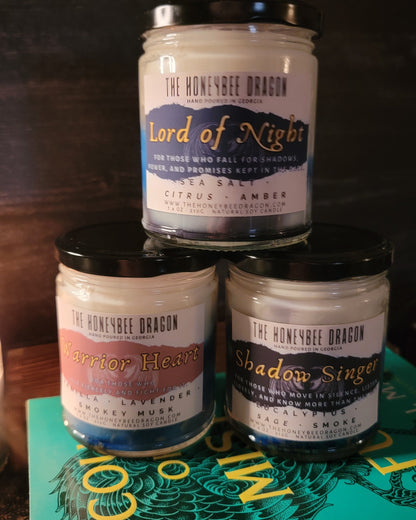 Three stacked soy candles from The HoneyBee Dragon: Lord of Night, Warrior Heart, and Shadowsinger, displayed on a fantasy book against a dark background.