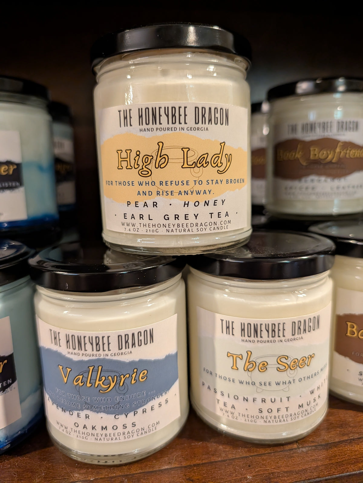 Three fantasy-inspired soy candles from The HoneyBee Dragon: High Lady, Valkyrie, and The Seer, arranged in a pyramid with other candles in the background.